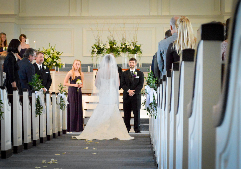 a bride walking down the aisle at a church with her groom in the background. Flower petals trail her on the ground. 