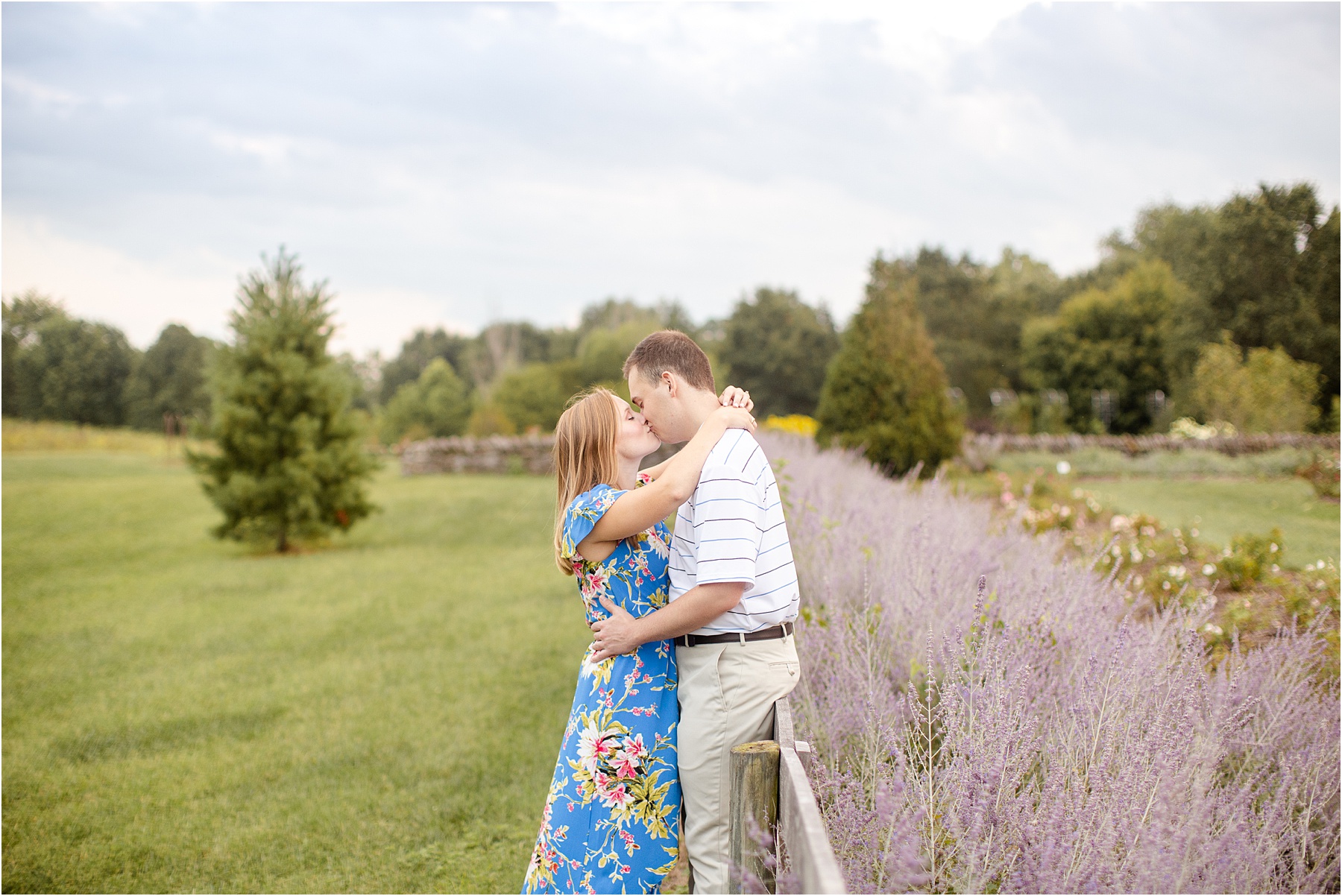 Man leans up against fence of lavender field for engagement pictures