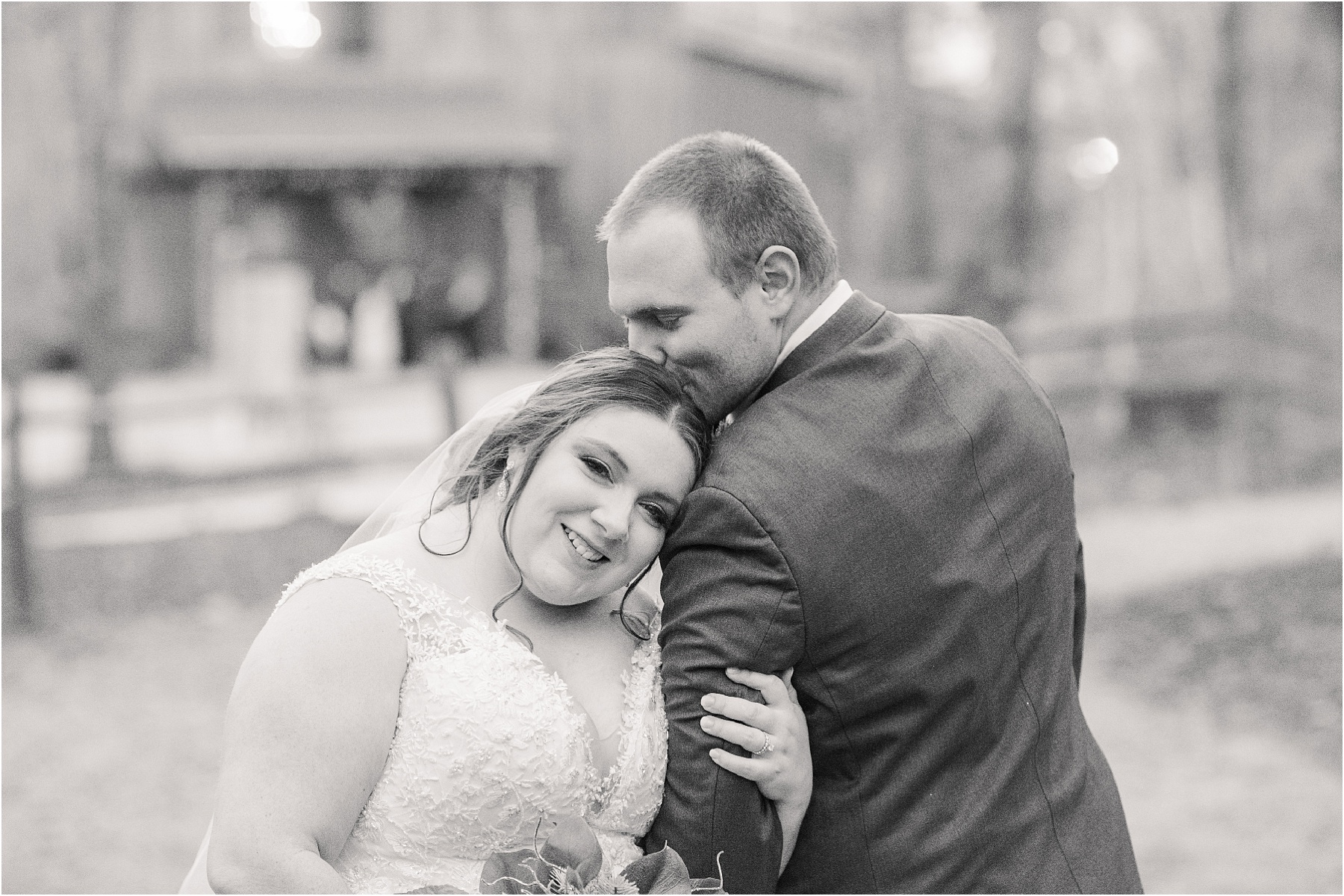black and white photo of a bride looking at the camera and groom kissing her head